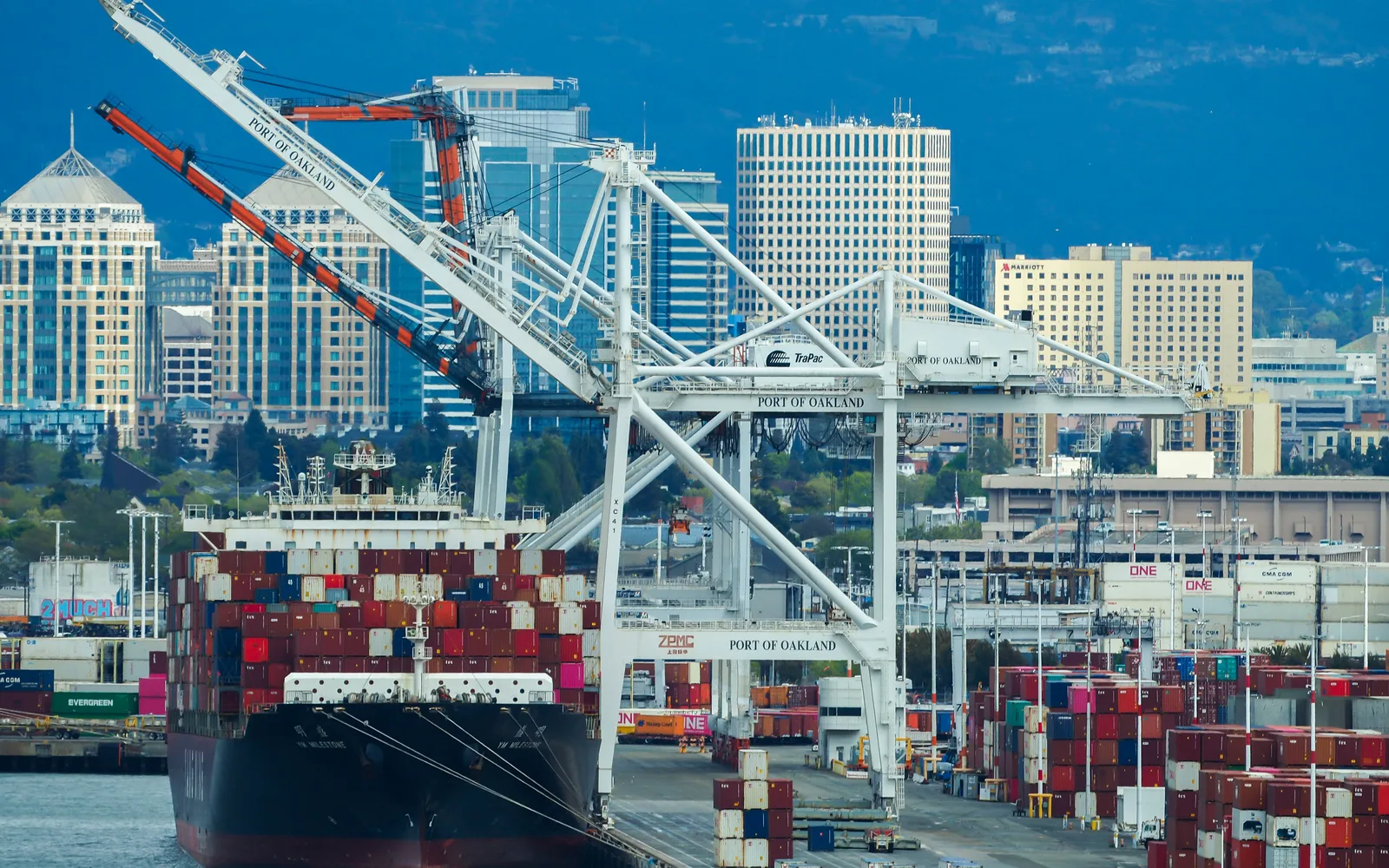 A container ship docked at the Port of Oakland with cargo cranes visible and the Oakland Hills in the background